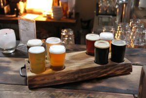 Eight beer samples in a burned wood holder on a burned wood table in a warm room.